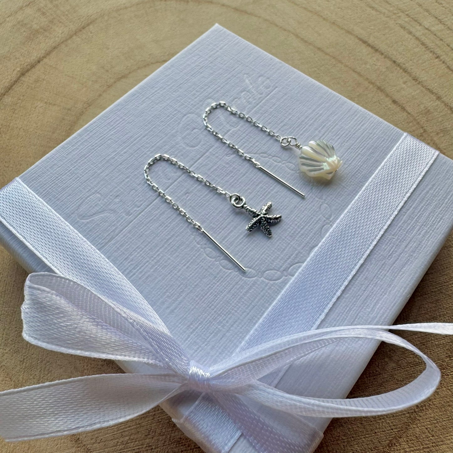 Pair of sterling silver earrings with starfish and shell designs on a white box with a white ribbon, placed on a wooden surface.