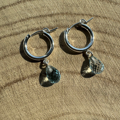 Sterling silver hoop earrings with teardrop-shaped blue topaz charms on a wooden surface