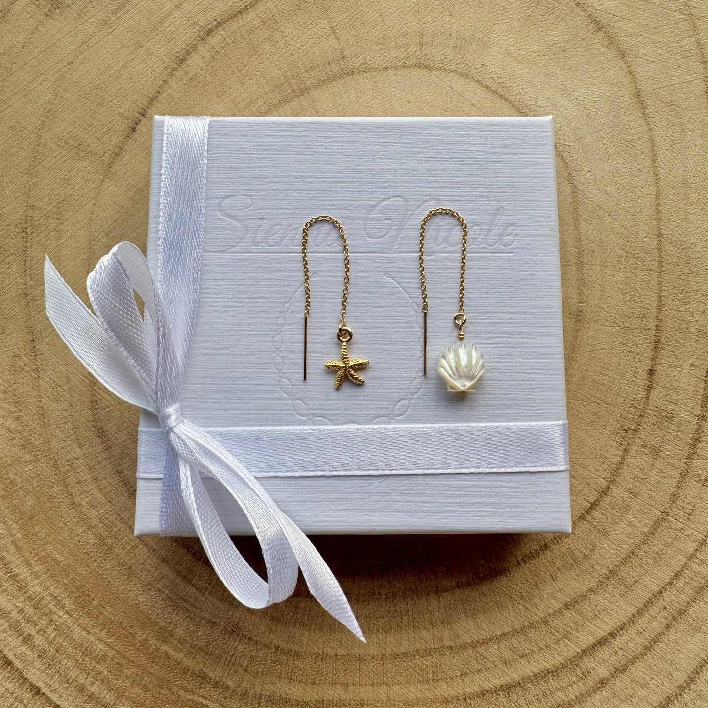 Pair of gold filled earrings with starfish and shell designs on a white box with a white ribbon, placed on a wooden surface.