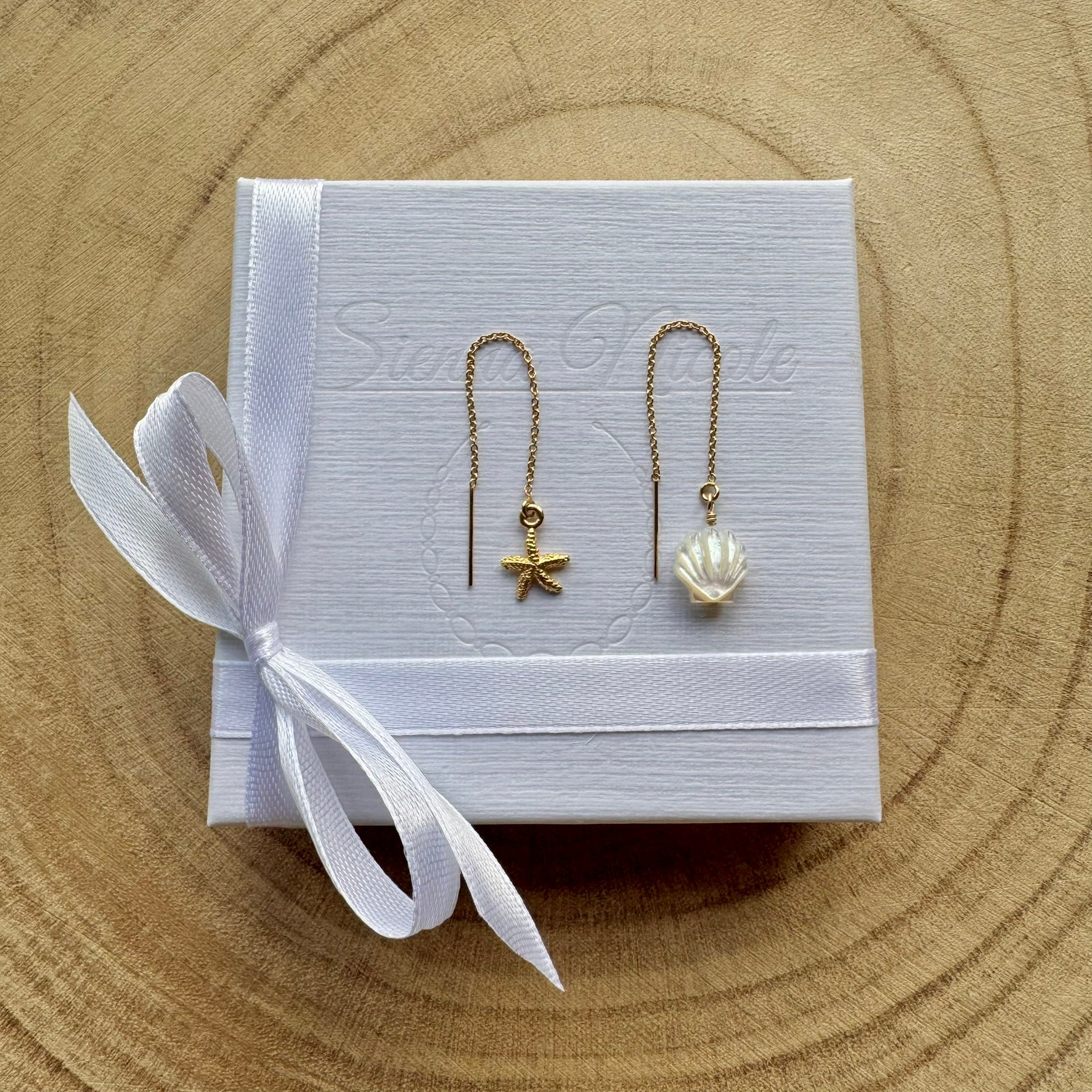 Pair of gold filled earrings with starfish and shell designs on a white box with a white ribbon, placed on a wooden surface.