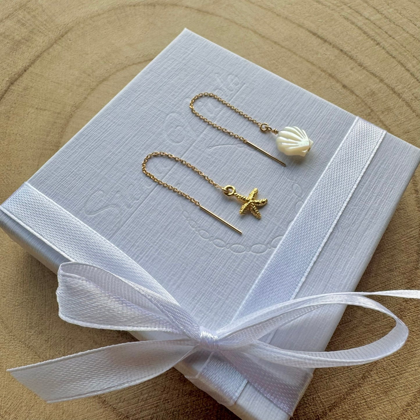 Pair of gold filled earrings with starfish and shell designs on a white box with a white ribbon, placed on a wooden surface.