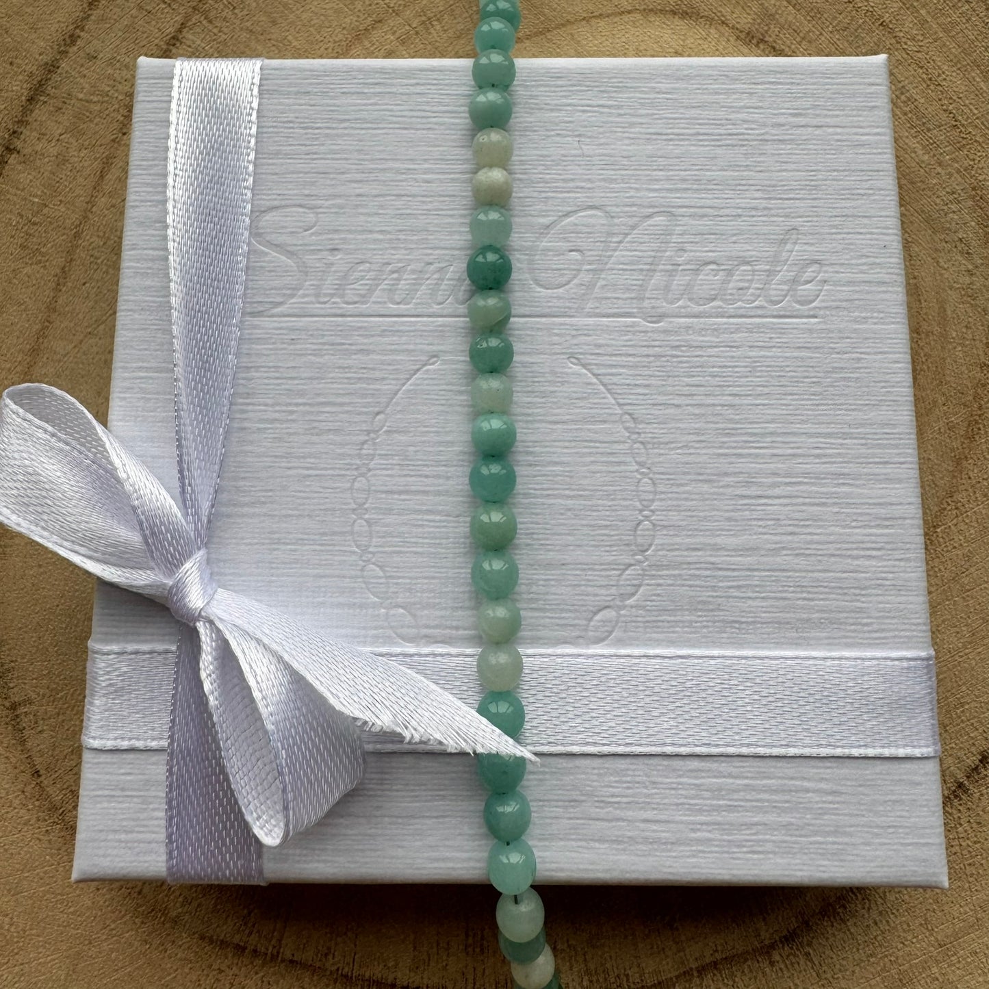 White gift box with a white ribbon and a blue green amazonite gemstone beaded bracelet on a wooden surface