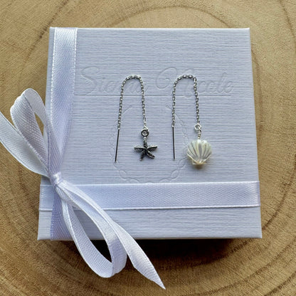 Pair of sterling silver earrings with starfish and shell designs on a white box with a white ribbon, placed on a wooden surface.