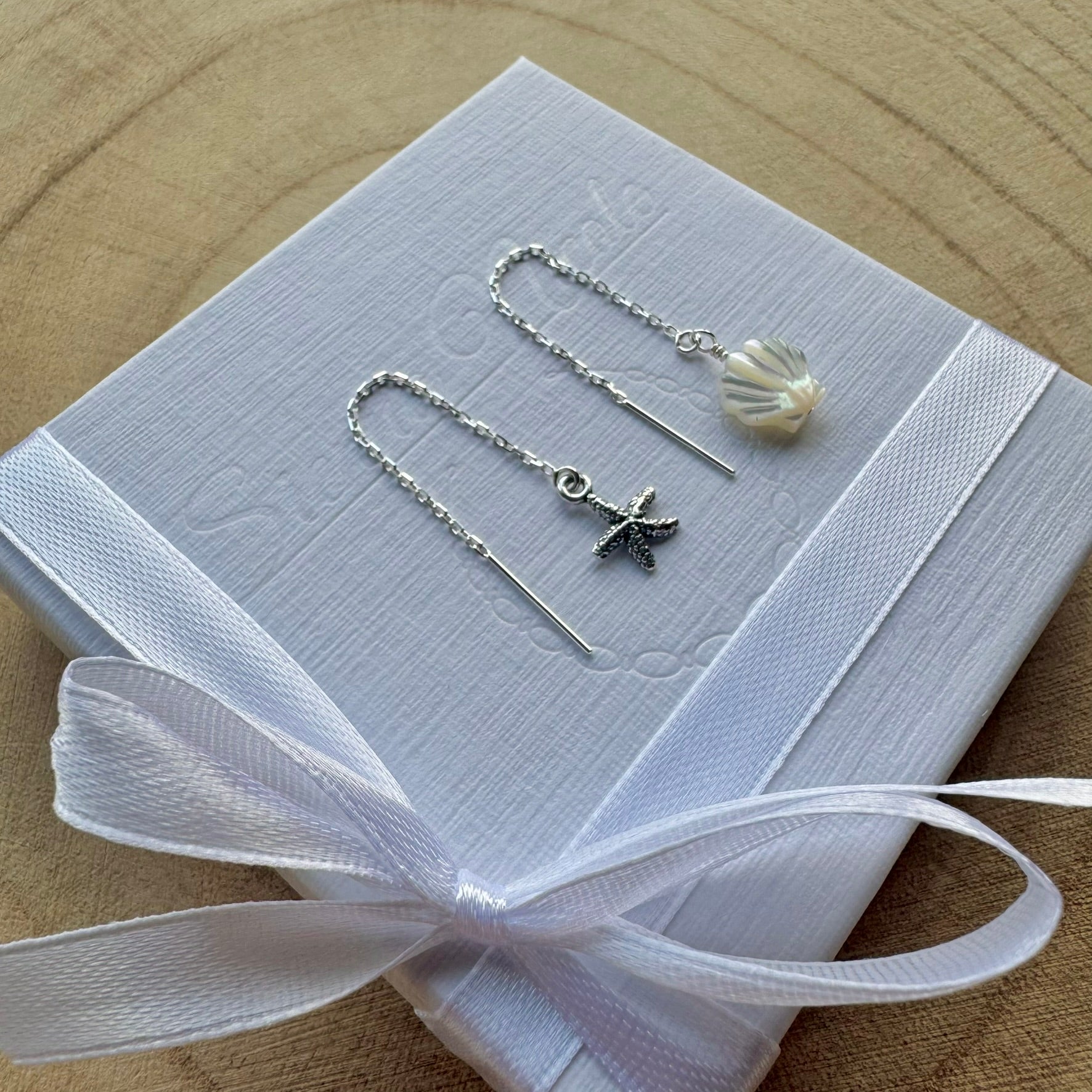 Pair of sterling silver earrings with starfish and shell designs on a white box with a white ribbon, placed on a wooden surface.